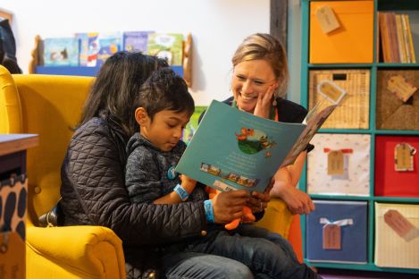 Mother sits with child on her lap reading a picture book with a smiling woman watching in the background